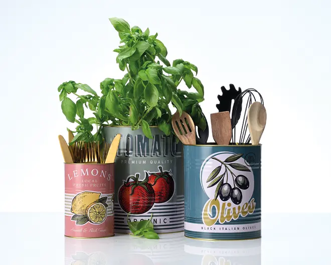 Tre cylindriske metaldåser med retro-inspirerede etiketter vises. Den første dåse, dekoreret med citroner, indeholder guldfarvet bestik. Den anden, større dåse med tomatmotiv, rummer en frodig grøn basilikumplante. Den tredje dåse, med olivendesign, er fyldt med forskellige køkkenredskaber såsom træskeer, et piskeris og en pastaske. Hver dåse har et farverigt, nostalgisk udtryk og bruges til praktisk opbevaring i køkkenet.