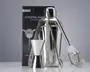 Et sæt barværktøj i rustfrit stål, herunder en cocktailshaker med blank overflade, en dobbeltsidet målebæger til væsker, en lang røreske, et par istænger og en cocktail-si med spiralformet fjeder. Produkterne er vist foran en mærket æske, og alle dele har et stilrent, moderne design, der passer til både hjemmebrug og professionel brug.