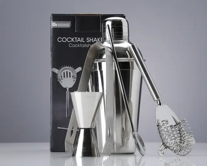 Et sæt barværktøj i rustfrit stål, herunder en cocktailshaker med blank overflade, en dobbeltsidet målebæger til væsker, en lang røreske, et par istænger og en cocktail-si med spiralformet fjeder. Produkterne er vist foran en mærket æske, og alle dele har et stilrent, moderne design, der passer til både hjemmebrug og professionel brug.