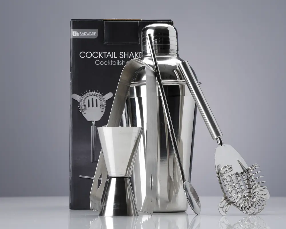 Et sæt barværktøj i rustfrit stål, herunder en cocktailshaker med blank overflade, en dobbeltsidet målebæger til væsker, en lang røreske, et par istænger og en cocktail-si med spiralformet fjeder. Produkterne er vist foran en mærket æske, og alle dele har et stilrent, moderne design, der passer til både hjemmebrug og professionel brug.