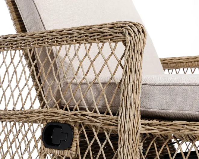 Nærbillede af en stol med ramme i flettet rattan og diamantformet gittermønster. Stolen har tykke, lyse beige hynder på sæde og ryg, som giver god komfort. På siden ses en sort plastikgreb eller drejeknap, sandsynligvis til at justere stolens position eller vippefunktion. Designet kombinerer naturlige materialer med moderne funktionalitet.