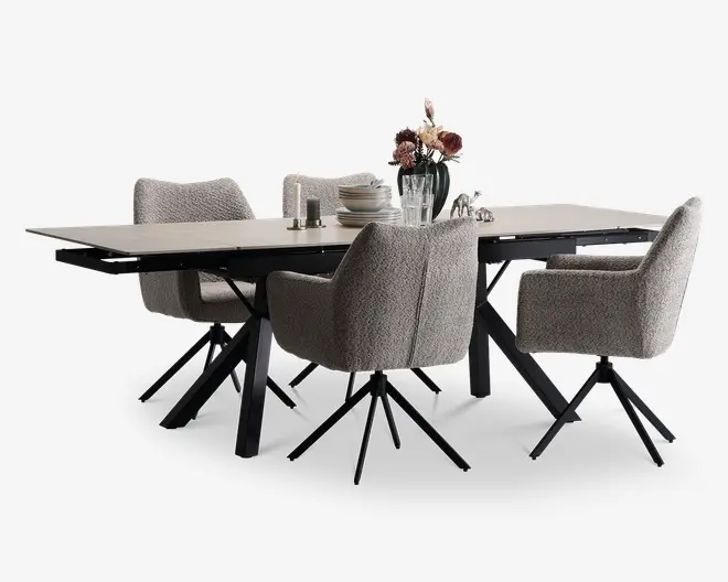 Et moderne spisesæt bestående af et rektangulært bord med en lys, glat bordplade og solide sorte metalben. Sættet inkluderer fire polstrede stole med blødt, tekstureret gråt stof, armlæn og sorte metalben med drejefunktion. Bordet er pyntet med en sort vase med lyserøde og hvide blomster, en stak hvide tallerkener, klare drikkeglas samt salt- og peberbøsser, hvilket skaber en indbydende og moderne spiseatmosfære.