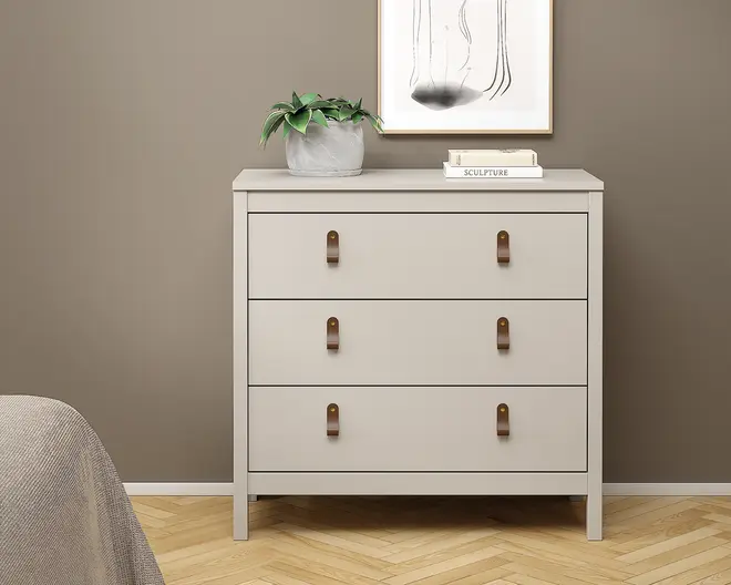Et moderne, minimalistisk møbel med tre rummelige skuffer, hver med to brune læderstropper som greb for nem åbning. Kommoden har en glat, mat lysebeige finish og står på korte, solide ben. Toppen er dekoreret med en lille potteplante og en stak bøger, hvilket giver et dekorativt præg. Det samlede design er enkelt og stilrent, velegnet til moderne indretning.