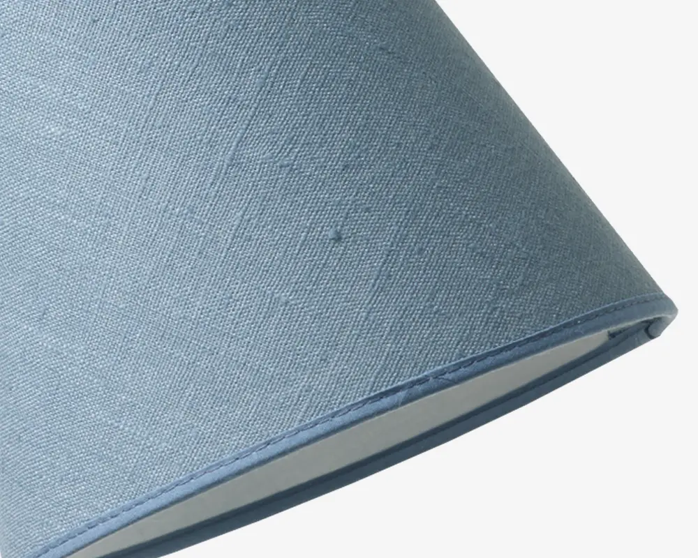 Nærbillede af en lampeskærm med klassisk konisk form, betrukket med lyseblåt tekstil med en let vævet struktur. Stoffet har en diskret mønster, og kanterne er pænt afsluttet med matchende blå kantbånd. Den indvendige foring er i en lys, neutral farve.