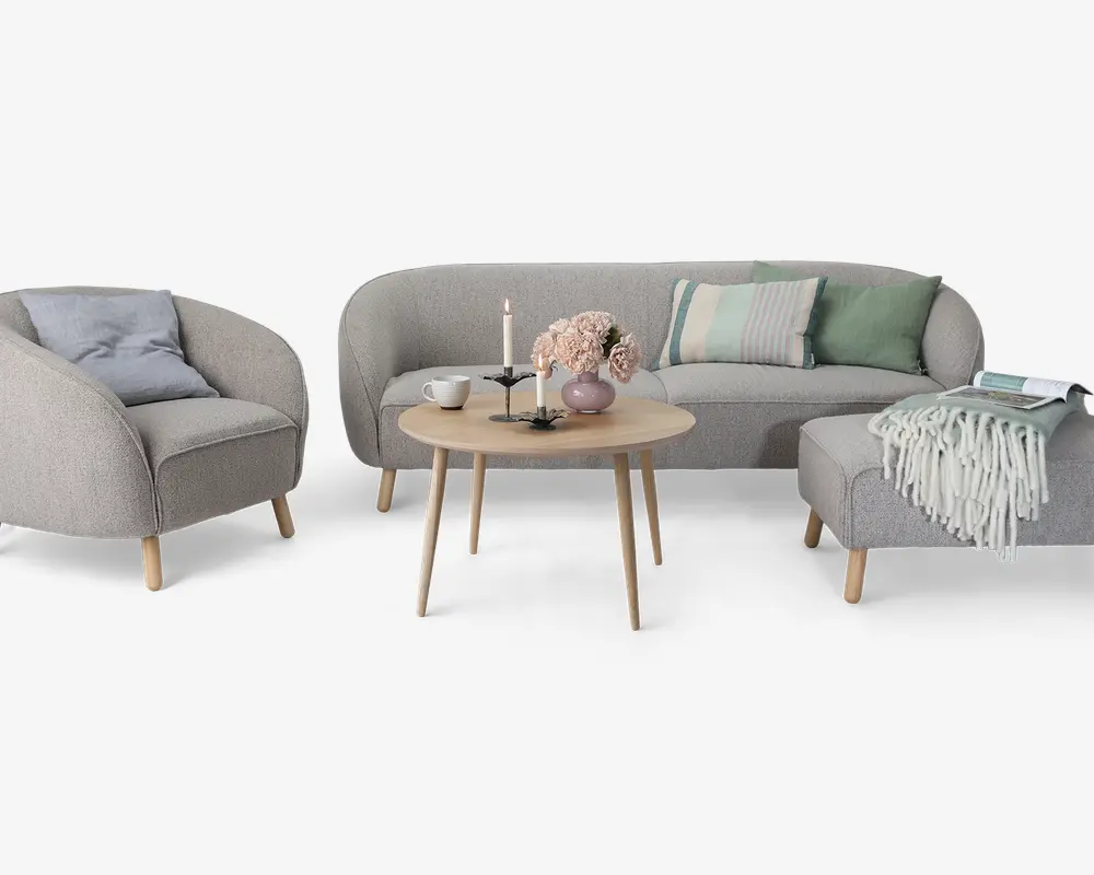 Et moderne stuesæt bestående af en lysegrå lænestol, en matchende buet sofa med pastelfarvede grønne og stribede puder samt en rektangulær skammel med et tykt strikket tæppe over. Sættet inkluderer et rundt sofabord i lyst træ, dekoreret med en vase med lyserøde blomster, to lysestager og en kop. Alle møbler har lyse træben, hvilket giver et skandinavisk inspireret udtryk.