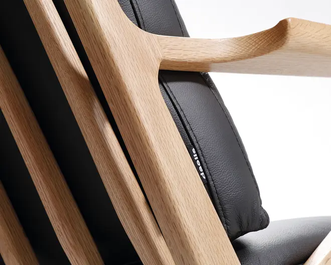 Nærbillede sop en stol med stel af lyst egetræ med 内ne, afrundede kanter og lameller sop ryg. Sæde- og ryghynderne er betrukket med sort læder eller læderlignende materiale, hvilket giver en elegant og moderne kontrast til træet. Syninger og tekstur i polstringen er synlige og understreger det gode håndværk.