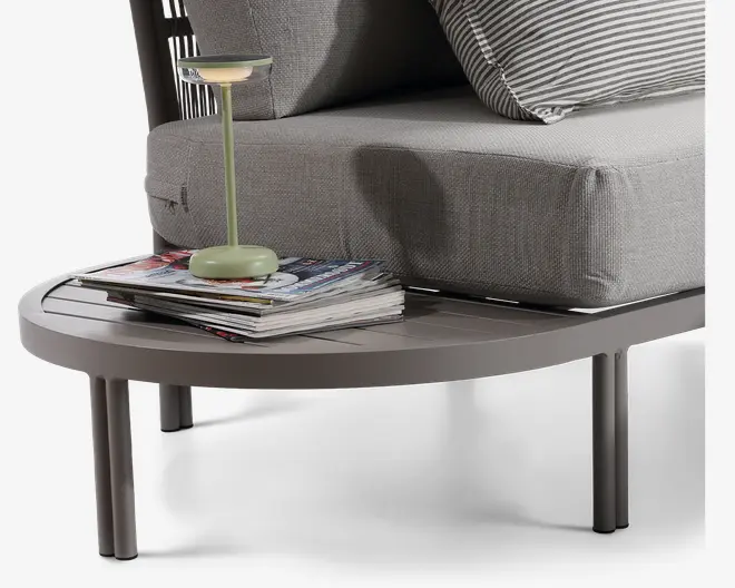 Nærbillede af et moderne, rundt sidebord med en glat, mat overflade i en neutral farve. Bordet har slanke, cylindriske ben og en overflade med lameller. På bordet står en lille, minimalistisk lampe med lysegrøn fod og stamme samt en stak magasiner. I baggrunden ses en del af en sofa med lysegråt, tekstureret betræk og en stribet pude.