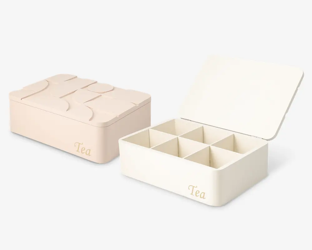 Rektangulær opbevaringsboks med hængslet låg, vist i to pastelfarver. Boksen har ordet 'Tea' trykt på forsiden og indeholder seks indvendige rum til organisering. Designet er minimalistisk og moderne med glatte kanter og en ren finish.