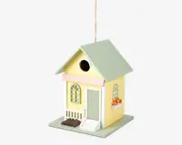 Dekorativt fuglehus formet som et lille gult hus med lysegrønt tag, der har en lille veranda med hvidt gelænder, grøn dør og buede vinduer. Der er en lille hvid trappe op til døren, en blomsterkasse med orange blomster under et vindue, og et rundt indgangshul over verandaen.