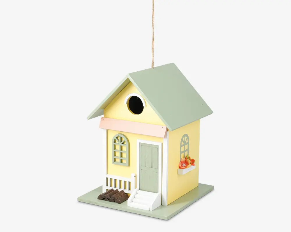 Dekorativt fuglehus formet som et lille gult hus med lysegrønt tag, der har en lille veranda med hvidt gelænder, grøn dør og buede vinduer. Der er en lille hvid trappe op til døren, en blomsterkasse med orange blomster under et vindue, og et rundt indgangshul over verandaen.