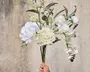 En hånd holder en buket med kunstige hvide blomster, herunder hortensia, orkidéer og kamelia. Buketten har realistiske grønne blade og små hvide fyldblomster samt nogle knopper og uåbnede blomster. Stilkene er lange og ser naturlige ud, og kronbladene har en blød, naturtro tekstur. Udtrykket er elegant og frisk, velegnet til dekoration.