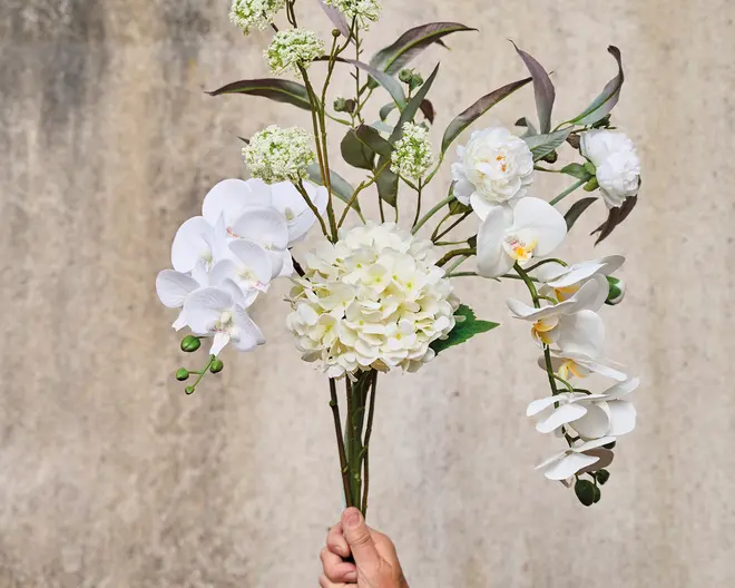 En hånd holder en buket med kunstige hvide blomster, herunder hortensia, orkidéer og kamelia. Buketten har realistiske grønne blade og små hvide fyldblomster samt nogle knopper og uåbnede blomster. Stilkene er lange og ser naturlige ud, og kronbladene har en blød, naturtro tekstur. Udtrykket er elegant og frisk, velegnet til dekoration.