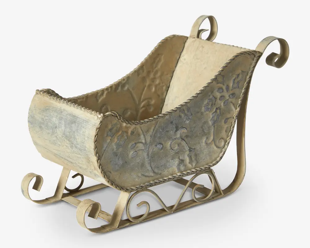 Dekorativ beholder formet som en slæde, fremstillet af metal med antik, patineret finish. Siderne har prægede blomster-mønstre og udsmykkede krøllede detaljer, mens bunden har buede meder og indviklede metaludsmykninger. Farven er en blanding af creme og dæmpet grå, hvilket giver et vintage udseende.