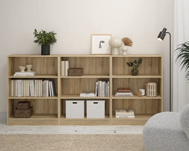 En reol i lyst egetræslook med tre sektioner, hver med to åbne hylder. Hylderne er dekoreret med en blanding af bøger, dekorative kurve, hvide opbevaringskasser og forskellige pyntegenstande som vaser, en indrammet plakat og en lille plante. Reolen har et moderne, minimalistisk design og står på et lyst gulv med en neutral væg i baggrunden.