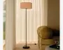 Høj gulvlampe med slank sort stang, rund fod og plisseret lyserød tromleformet skærm, vist ved siden af en sofa i fløjl i et blødt oplyst rum med transparente gardiner.