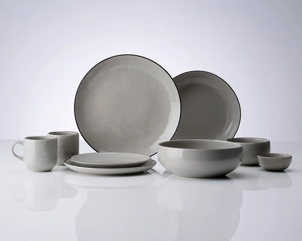 Et sæt stentøj i en blød grå farve med en let ujævn glasur. Sættet indeholder store middagstallerkener, mindre frokosttallerkener, dybe skåle, en lille skål og krus. Hver del har et minimalistisk design med glatte, afrundede kanter og en tynd, mørkere kant, der fremhæver formen. Udtrykket er moderne og enkelt, velegnet til både hverdags- og festbrug.