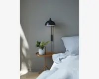 Et moderne soveværelsesmiljø med et natbord i lyst træ med rund bordplade og trefod. På bordet står en hvid keramisk vase med grønne blade. Ved siden af bordet er en seng med blåt og hvidt stribet sengetøj og puder. Over bordet hænger en sort væglampe med kuppelformet skærm. Væggen er malet i en blød grå farve, hvilket skaber en rolig og minimalistisk stemning.