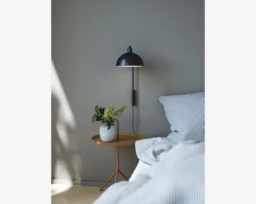 Et moderne soveværelsesmiljø med et natbord i lyst træ med rund bordplade og trefod. På bordet står en hvid keramisk vase med grønne blade. Ved siden af bordet er en seng med blåt og hvidt stribet sengetøj og puder. Over bordet hænger en sort væglampe med kuppelformet skærm. Væggen er malet i en blød grå farve, hvilket skaber en rolig og minimalistisk stemning.