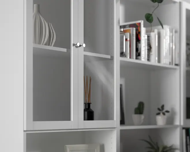 Nærbillede af en hvid reol med glaslåger og runde sølvfarvede greb. Indenfor på hylderne står dekorative genstande, herunder en moderne hvid vase med bølget design og en sort duftspreder med pinde. De tilstødende åbne hylder indeholder bøger, indrammede billeder og små potteplanter, hvilket giver reolen et rent og organiseret udtryk.