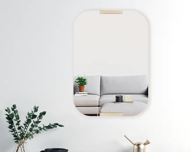 Rektangulært vægspejl med afrundede hjørner, udstyret med to guldfarvede metalbeslag øverst og nederst til ophængning. Spejlet har et minimalistisk design og reflekterer en moderne stue med en lysegrå hjørnesofa, en lille potteplante oven på en stak bøger samt et sofabord med et stearinlys og en bog. Spejlets enkle linjer og diskrete detaljer gør det velegnet til moderne indretning.