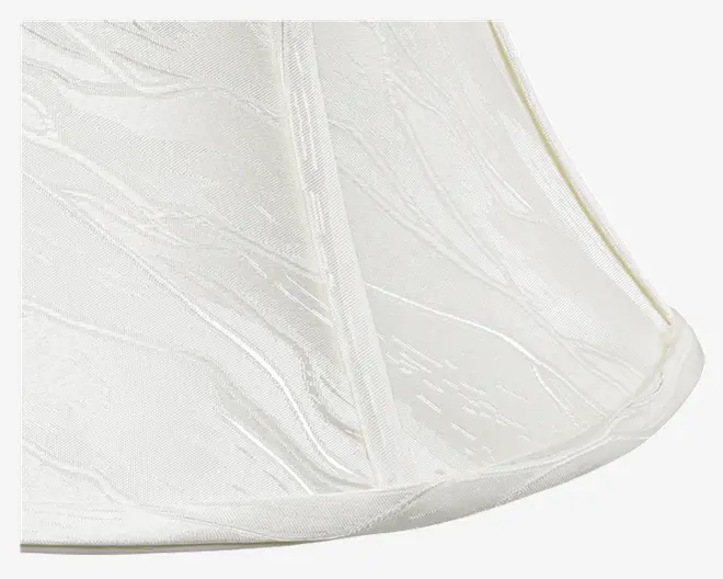 Nærbillede af en lampeskærm med blødt hvidt stof og et diskret tekstureret mønster. Stoffet har bløde, bølgende linjer og en let glans, hvilket giver et elegant udtryk. Lampeskærmen har en klassisk, udvidet form med forstærkede lodrette sømme for stabilitet.
