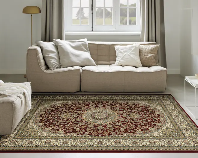 Rektangulært tæppe med et indviklet blomster-medaljonmønster i midten, med dybe røde, beige og gyldne nuancer. Tæppets kant er dekoreret med detaljerede motiver i matchende farver, hvilket giver et klassisk og elegant udtryk. Tæppet har lav luv og ser blødt ud, velegnet til brug i stuen.