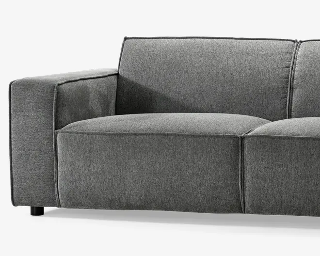 Denne vare har et moderne, minimalistisk design med rene linjer og en firkantet form. Den er betrukket med et mellemgråt stof, der virker blødt og slidstærkt. Sæde- og ryghynderne er tykke og bløde, hvilket giver god komfort. Armlænet er bredt og polstret, og matcher den samlede struktur. Syningerne er synlige og tilføjer en diskret detalje langs kanterne. Benene er korte og sorte, hvilket giver møblet en solid base.