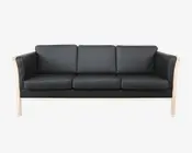 En moderne tresæders med sort læderbetræk, der har tre sædehynder og tre ryghynder. Stellet er lavet af lyst træ med en naturlig finish, som er synligt langs armlæn og bund. Armlænene er lige og let udadbuede, hvilket giver et moderne udtryk. Sæde- og ryghynderne er tykke og firkantede, hvilket giver et struktureret og støttende udseende.