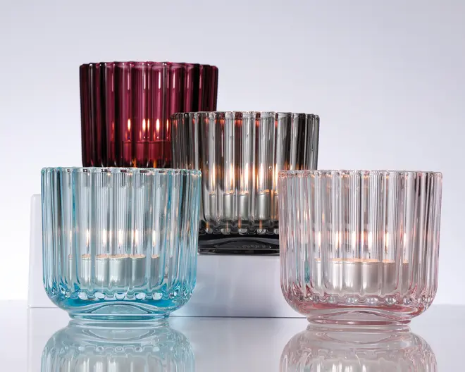 Sæt med fire rillede fyrfadsstager i glas i forskellige farver: dyb bordeaux, røggrå, lyseblå og sart rosa. Hver stage har en cylindrisk form med lodrette riller og en kraftig, solid bund. Glasset er halvgennemsigtigt, hvilket lader lyset fra fyrfadslyset sprede sig blødt gennem den strukturerede overflade og skaber en varm og indbydende glød. Stagerne er placeret i forskellige højder for at fremhæve farver og design.