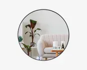 Rundt vægspejl med en tynd sort ramme, der reflekterer en moderne stue. I spejlet ses en lys beige, polstret sofa med buede armlæn, et træsofabord med dekorative genstande, herunder et hvidt lys, en lille plante og en blok-kalender. Der er også en høj potteplante med brede grønne og rødlige blade samt en hvid væg i baggrunden.