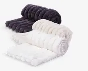 Tre bløde, sammenrullede tæpper vises på række, hver med et ribbet, tekstureret design. Tæpperne fås i tre farver: mørkegrå, creme og hvid. Materialet ser meget blødt og tykt ud, hvilket giver en hyggelig og luksuriøs fornemmelse. Kanterne er pænt afsluttede, og tæpperne har en let blank overflade, hvilket indikerer et højkvalitets, pelslignende stof.