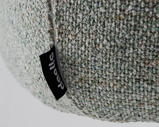 Nærbillede af en tekstureret, vævet stofoverflade i lysegrå med små prikker af blå, grøn og gul. Et sort stofmærke med hvid tekst er syet ind i en søm og viser mærket. Stoffet ser tykt og slidstærkt ud, velegnet til polstring eller boligindretning.