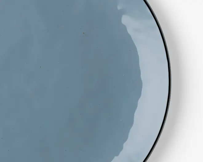 Et nærbillede af en rund, lyseblå keramiktallerken med blank finish. Tallerkenen har en glat overflade og en let hævet kant. Glasuren giver subtile farvevariationer og en reflekterende glans, hvilket giver tallerkenen et moderne og elegant udseende.