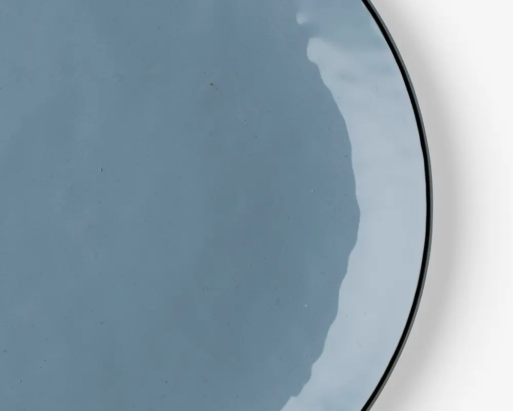 Et nærbillede af en rund, lyseblå keramiktallerken med blank finish. Tallerkenen har en glat overflade og en let hævet kant. Glasuren giver subtile farvevariationer og en reflekterende glans, hvilket giver tallerkenen et moderne og elegant udseende.