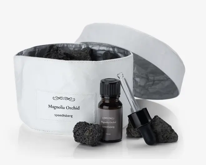 Et sæt bestående af en hvid stofbeholder med et mærke, hvor der står 'Magnolia Orchid speeditsberg', fyldt med flere porøse sorte sten. Foran beholderen står en lille mørk glasflaske med sort skruelåg, også mærket 'Magnolia Orchid speeditsberg 10ml'. Ved siden af flasken ligger en separat glaspipette med sort gummiballon, og der er yderligere sorte sten placeret omkring flasken og pipetten. Beholderen har et matchende hvidt låg, som vises åbent og ligger bagved.