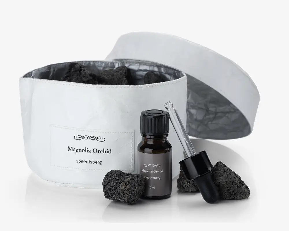 Et sæt bestående af en hvid stofbeholder med et mærke, hvor der står 'Magnolia Orchid speeditsberg', fyldt med flere porøse sorte sten. Foran beholderen står en lille mørk glasflaske med sort skruelåg, også mærket 'Magnolia Orchid speeditsberg 10ml'. Ved siden af flasken ligger en separat glaspipette med sort gummiballon, og der er yderligere sorte sten placeret omkring flasken og pipetten. Beholderen har et matchende hvidt låg, som vises åbent og ligger bagved.