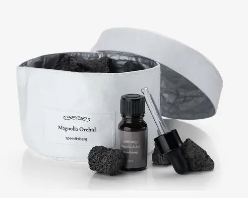 Et sæt bestående af en hvid stofbeholder med et mærke, hvor der står 'Magnolia Orchid speeditsberg', fyldt med flere porøse sorte sten. Foran beholderen står en lille mørk glasflaske med sort skruelåg, også mærket 'Magnolia Orchid speeditsberg 10ml'. Ved siden af flasken ligger en separat glaspipette med sort gummiballon, og der er yderligere sorte sten placeret omkring flasken og pipetten. Beholderen har et matchende hvidt låg, som vises åbent og ligger bagved.