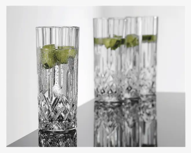Højt, klart glas med facetslebet diamantmønster i bunden, fyldt med en brusende drik og isterninger, pyntet med limebåde. Glasset har lodrette riller øverst og står sammen med tre lignende glas i baggrunden.