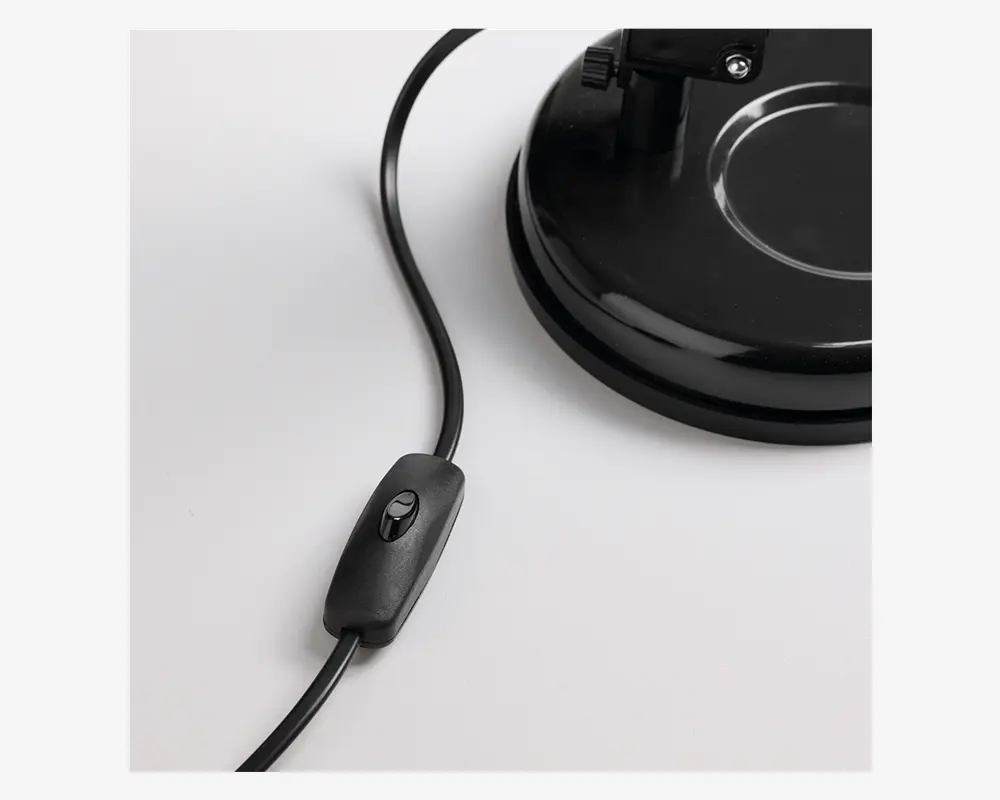 Nærbillede af en sort elektrisk ledning med en indbygget vippekontakt, der er forbundet til en robust, rund sort fod. Kontakten er nem at betjene med et enkelt tryk, og foden har en glat, blank overflade med en cirkulær fordybning for stabilitet.