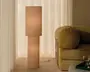 Høj gulvlampe med beige cylindrisk skærm og matchende fod, der giver et varmt og blødt lys. Stillet ved siden af en fløjlssofa i et roligt rum med neutrale farver og lette gardiner.