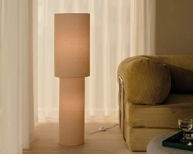 Høj gulvlampe med beige cylindrisk skærm og matchende fod, der giver et varmt og blødt lys. Stillet ved siden af en fløjlssofa i et roligt rum med neutrale farver og lette gardiner.