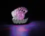En dekorativ genstand formet som en åben muslingeskal, lavet af skinnende metal. Inde i skallen ligger en stor, rund kugle fyldt med glimmer, der glitrer i lyserøde og lilla nuancer. Genstanden reflekterer og kaster farverige lysmønstre på overfladen nedenunder, hvilket skaber en livlig og iøjnefaldende effekt.