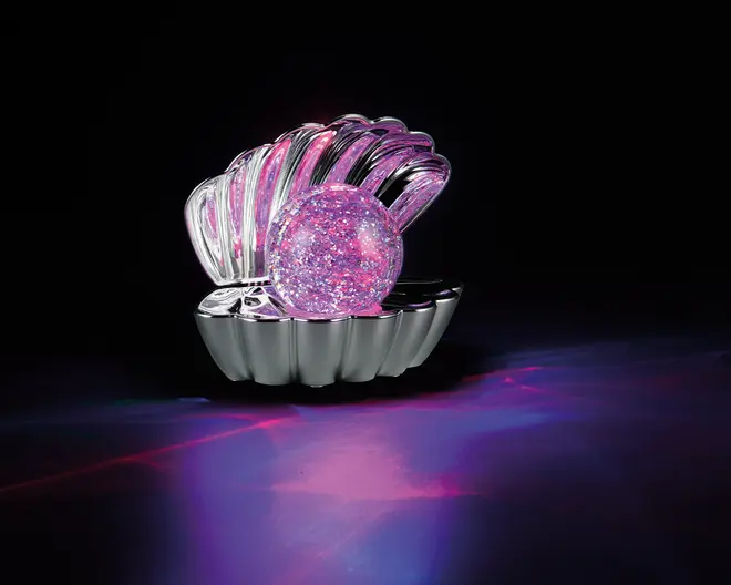 En dekorativ genstand formet som en åben muslingeskal, lavet af skinnende metal. Inde i skallen ligger en stor, rund kugle fyldt med glimmer, der glitrer i lyserøde og lilla nuancer. Genstanden reflekterer og kaster farverige lysmønstre på overfladen nedenunder, hvilket skaber en livlig og iøjnefaldende effekt.