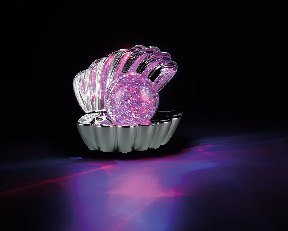 En dekorativ genstand formet som en åben muslingeskal, lavet af skinnende metal. Inde i skallen ligger en stor, rund kugle fyldt med glimmer, der glitrer i lyserøde og lilla nuancer. Genstanden reflekterer og kaster farverige lysmønstre på overfladen nedenunder, hvilket skaber en livlig og iøjnefaldende effekt.