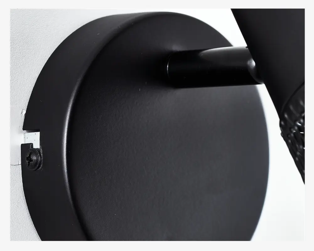 Nærbillede af et mat sort, cirkulært vægbeslag med glat overflade, synlig skrue i venstre side og et cylindrisk arm, der stikker ud. Designet er minimalistisk og moderne med rene linjer og et solidt udtryk.