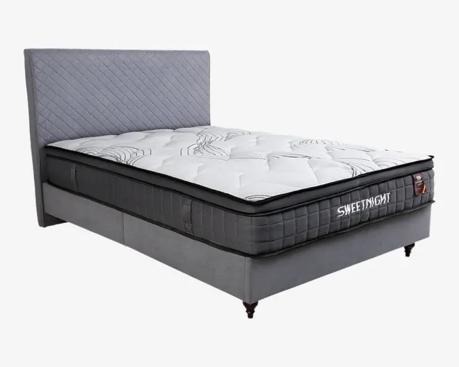 En moderne seng med et højt, quiltet gråt sengegærde og en tyk, blød madras. Madrassen har en hvid overflade med diskrete bølgemønstre og en mørkegrå, tekstureret side. Sengens bund er polstret med matchende gråt stof og står på korte, mørke ben. Designet er moderne, og materialerne ser bløde og komfortable ud.