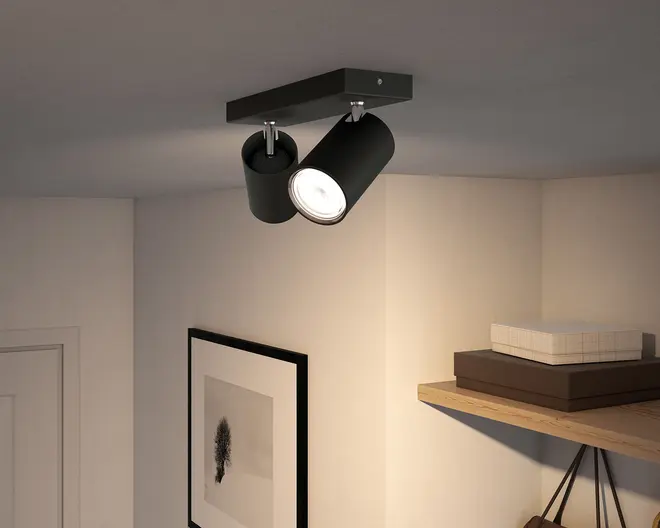 Sort, rektangulær loftmonteret spotlight med to justerbare cylindriske hoveder, hvor det ene peger nedad og er tændt, vist mod en neutral loft- og vægflade.
