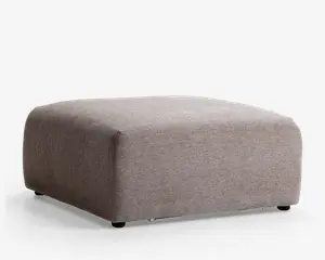 Firkantet puf med minimalistisk design, polstret med lysebrunt stof. Overfladen er glat, og hjørnerne er blødt afrundede, hvilket giver et moderne og alsidigt udtryk. Puffen står lavt på gulvet og har ingen synlige ben eller dekorative detaljer.