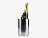 En høj, cylindrisk vinkøler i rustfrit stål med en blank, reflekterende overflade og lodrette riller langs siderne. Køleren indeholder en flaske champagne, hvor halsen og den guldfolierede top er synlig over kanten. Designet er stilrent og moderne, velegnet til at holde vin eller champagne kold.