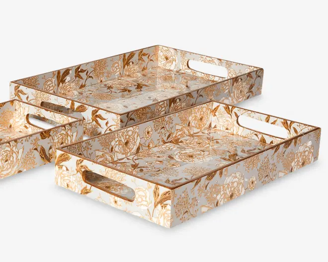 Sæt med tre rektangulære bakker i forskellige størrelser, hver med høje kanter og udskårne håndtag på de korte sider for nem transport. Bakkerne har en blank overflade og er dekoreret med et detaljeret blomstermønster i gyldne og beige nuancer på hvid baggrund, hvilket giver dem et elegant og dekorativt udtryk.